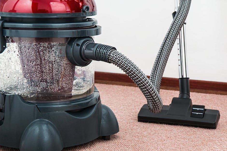 A person is kneeling on a patterned carpet in a residential interior, preparing a yellow and black portable vacuum cleaner for surface cleaning. The individual has curly hair and is wearing a khaki ja