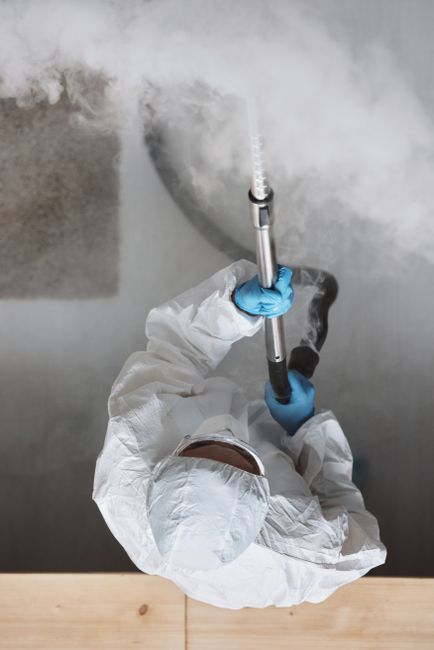 A professional cleaner wearing a white protective suit, blue gloves, and a face mask is operating a steam cleaning machine to sanitize a large, grey wall surface. The wall appears to have some grime o
