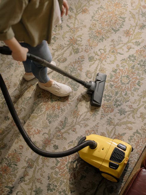 A professional cleaner wearing a white protective suit, blue gloves, and a face mask is operating a steam cleaning machine to sanitize a large, grey wall surface. The wall appears to have some grime o